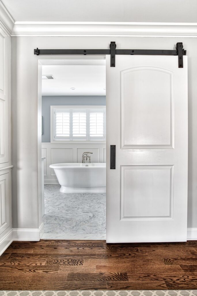 Spa-like bathroom with a white sliding barn door opening to a freestanding soaking tub, marble flooring, and soft gray walls. A serene wellness retreat atmosphere enhanced by natural light and clean architectural lines.