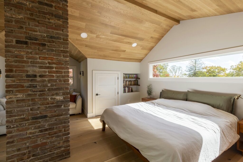 Home remodeling projects in Morris County: Modern attic primary bedroom with sloped wood ceiling, exposed brick column, and large horizontal window bringing in natural light. Features minimalist decor, light oak flooring, and a cozy reading nook.