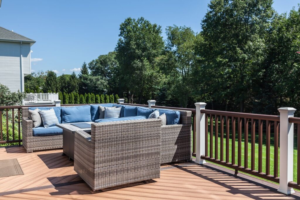 Spacious backyard deck with woven rattan sectional seating, blue cushions, and striped throw pillows surrounding a fire pit table. Features wood-look composite decking and a white-and-brown railing overlooking a green lawn and trees.