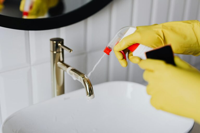 Person cleaning a bathroom faucet with spray cleaner and gloves to maintain fixture condition