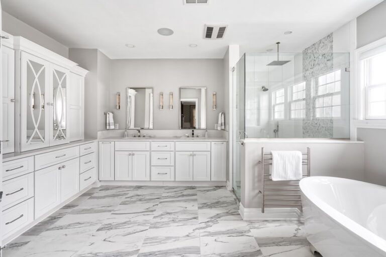 Elegant master bathroom renovation with marble-look tile, white cabinetry, glass-enclosed shower, and freestanding tub — featured by WarmlyYours.