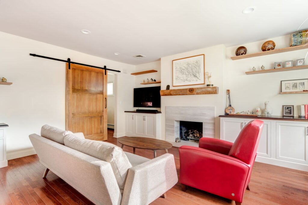 Living room interior after home remodeling with modern fireplace, built-in cabinets, floating shelves, sliding barn door, beige sofa, and red armchair on hardwood floors - Kraftmaster Renovations NJ