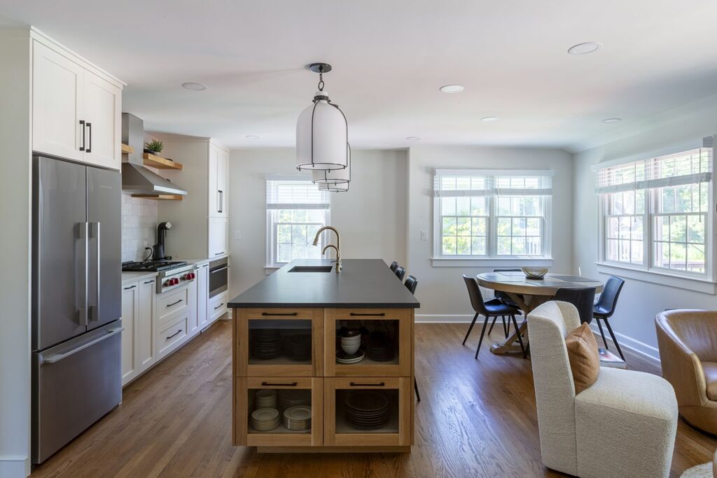 Bright modern kitchen with white cabinetry, stainless steel appliances, a large island with wood shelving and a dark countertop, pendant lighting, and an adjacent dining nook with large windows in Chatham NJ.
