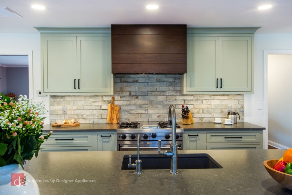 Sage green kitchen cabinets with dark wood hood, brick-style backsplash, and matte black countertops with stainless steel appliances and fixtures.